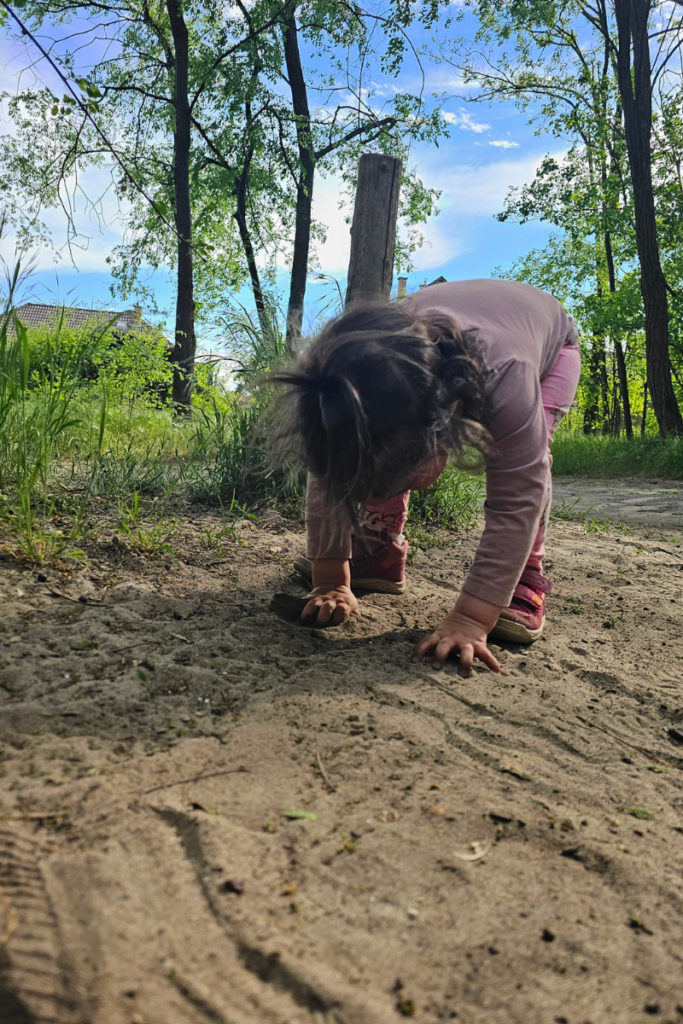 A toddler leaning down to touch sandy soil on a forest path, pressing both hands into the ground as a natural toddler sensory activity.
