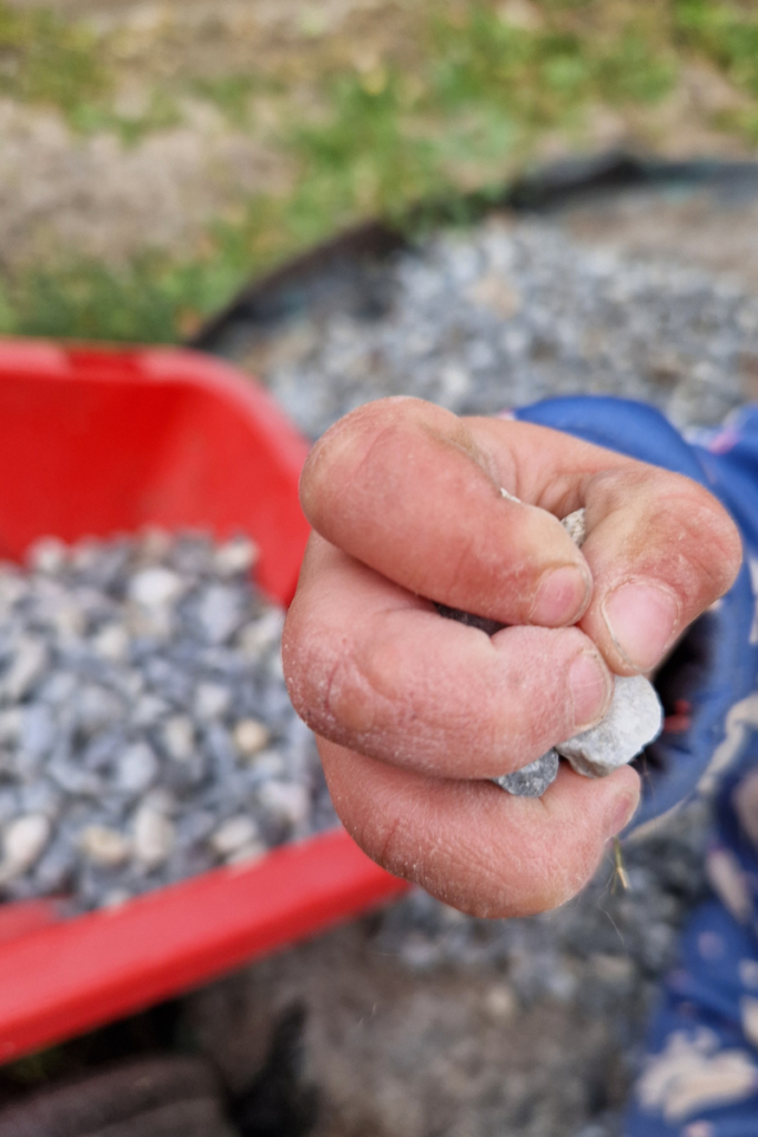 A toddler's small fist clutching a handful of small stones next to a red bucket, exploring texture and weight through hands-on outdoor play.
