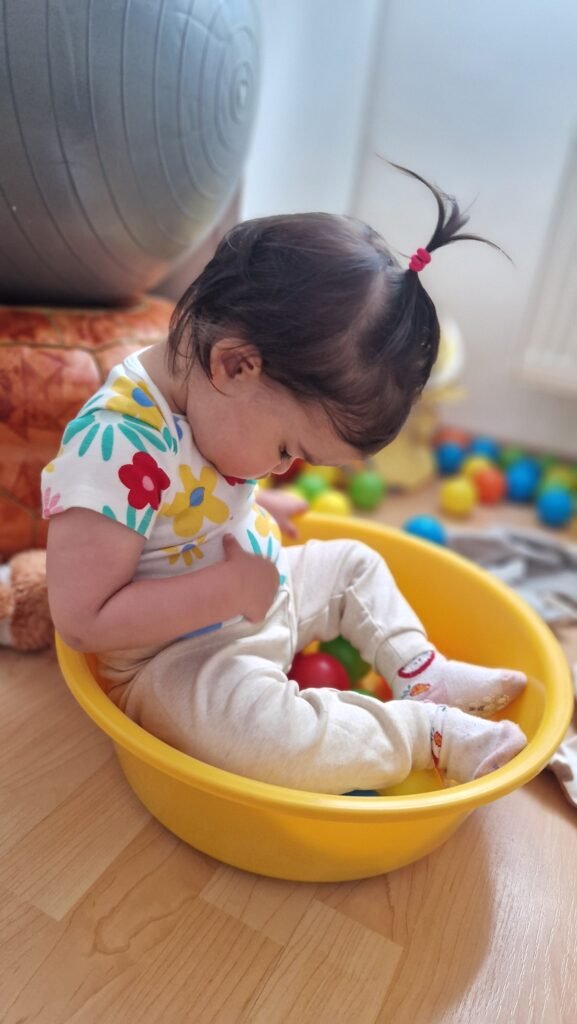 A toddler sitting inside a yellow plastic bowl on the floor, completely absorbed and satisfied — a perfect example of how everyday household items become the best toddler learning activities at home.