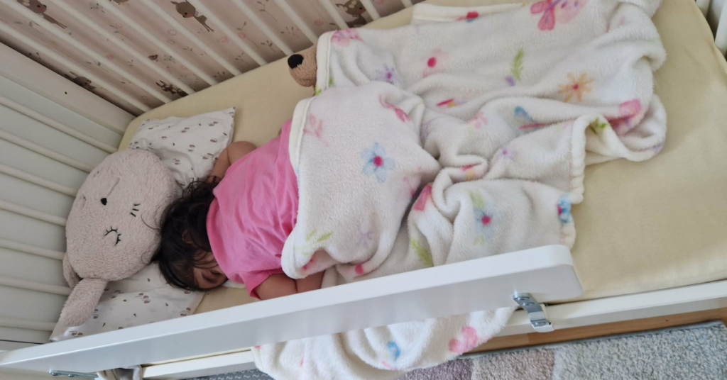 A toddler sleeping face-down in a cot surrounded by soft toys and a floral blanket, a peaceful moment that captures the quiet world of toddler sleep and the reality of toddler night terrors