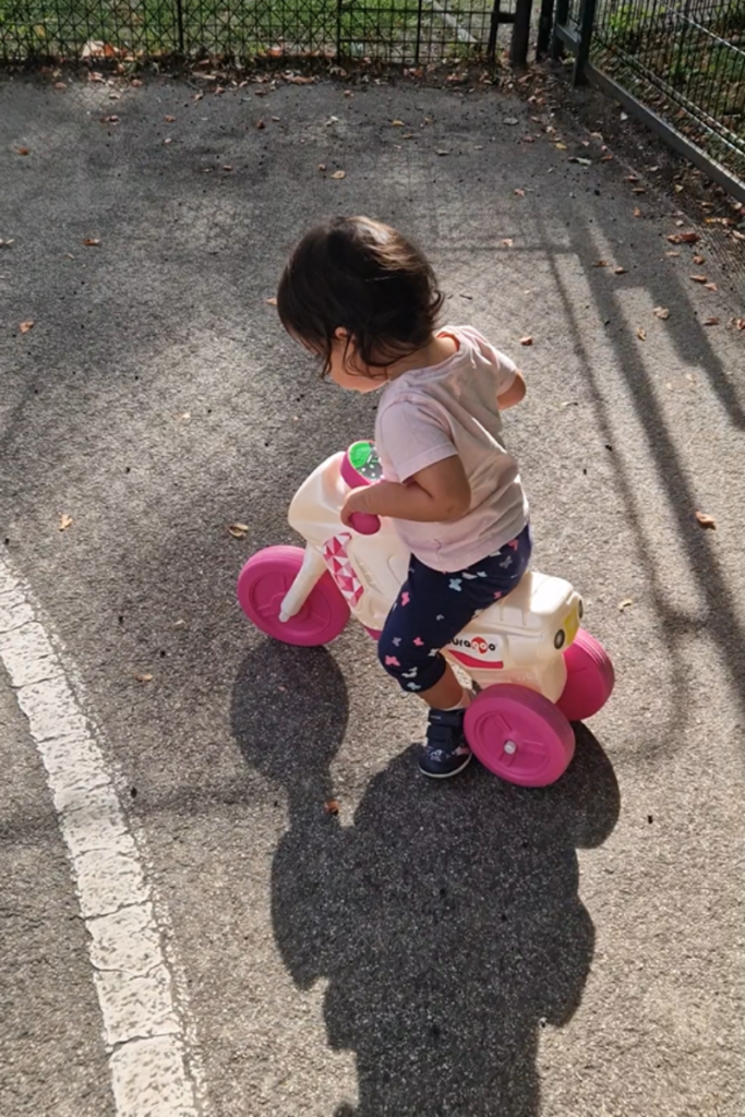 A toddler riding a white balance bike with pink wheels and handlebars on a playground, viewed from above, focused and determined — one of the best toddler learning activities for building coordination and persistence.