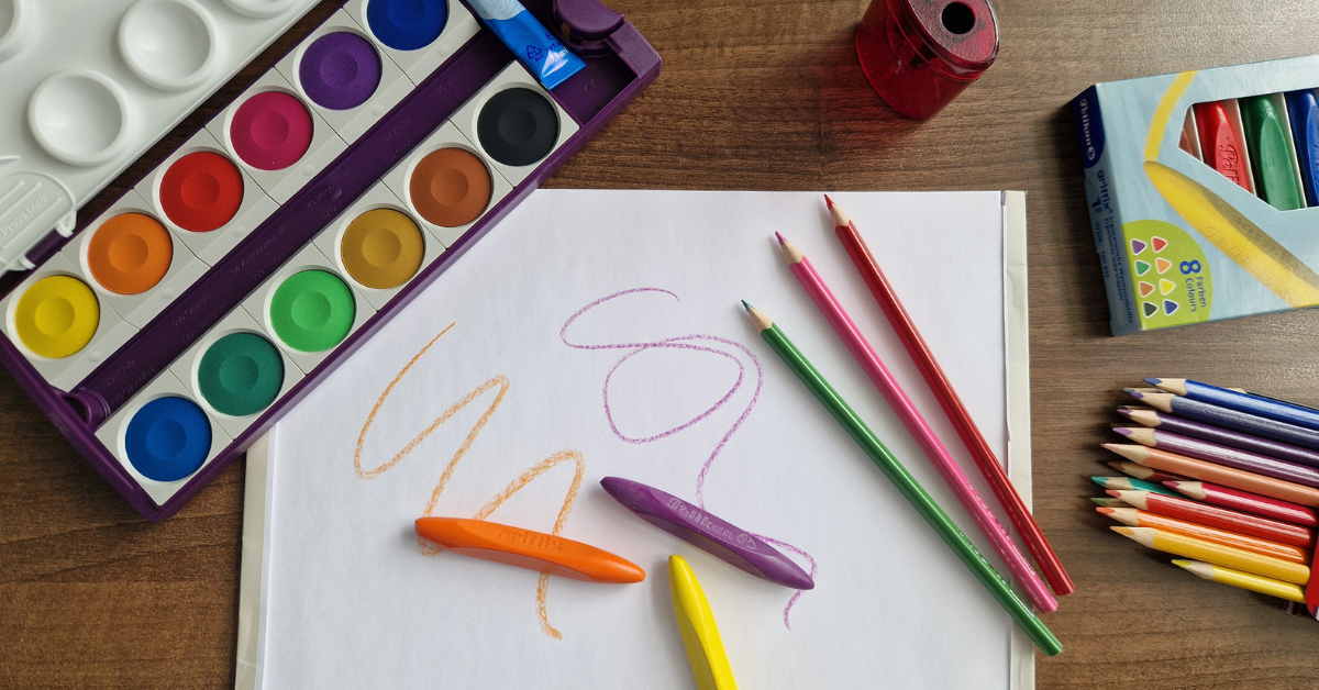 Main elementary school supplies list items laid out on a desk including watercolor paints, crayons, and colored pencils, chosen by a stationery professional and mom