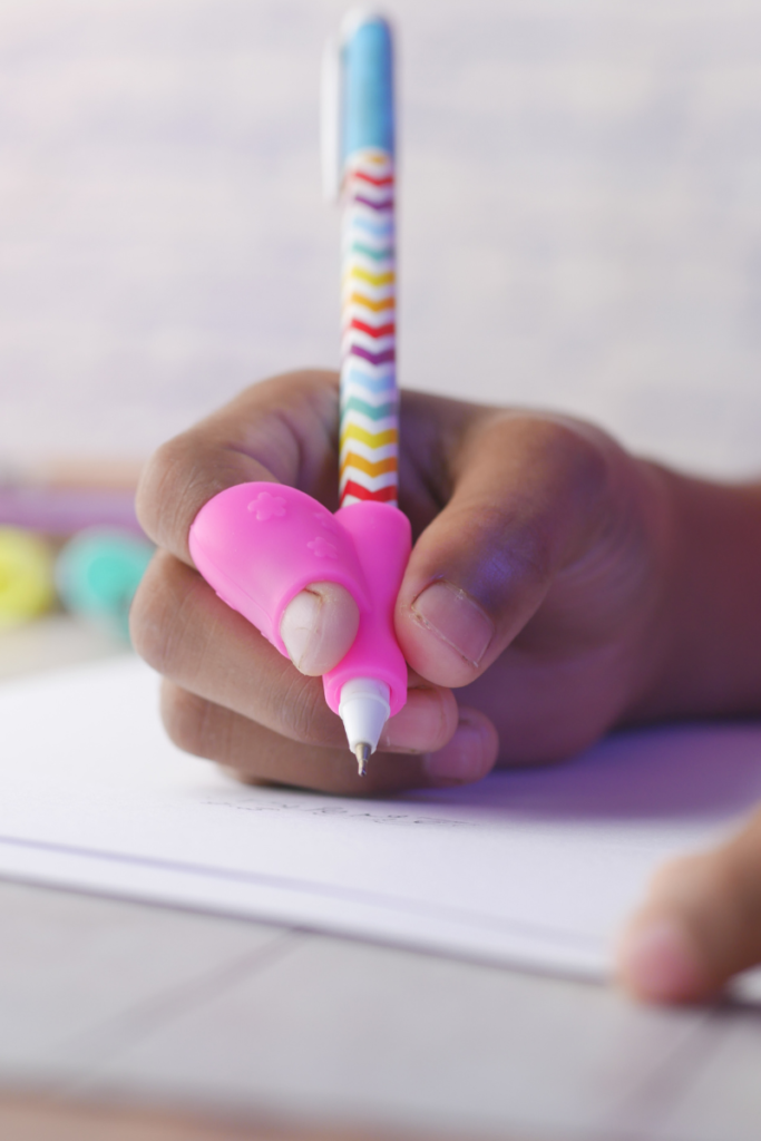 A child's hand writing with a pink molded pencil grip attached to a pen, one of the fine motor tools that guides finger placement