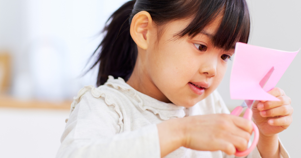 A young girl concentrating as she cuts a piece of pink paper with scissors, one of the everyday classroom tasks that depend on fine motor skills