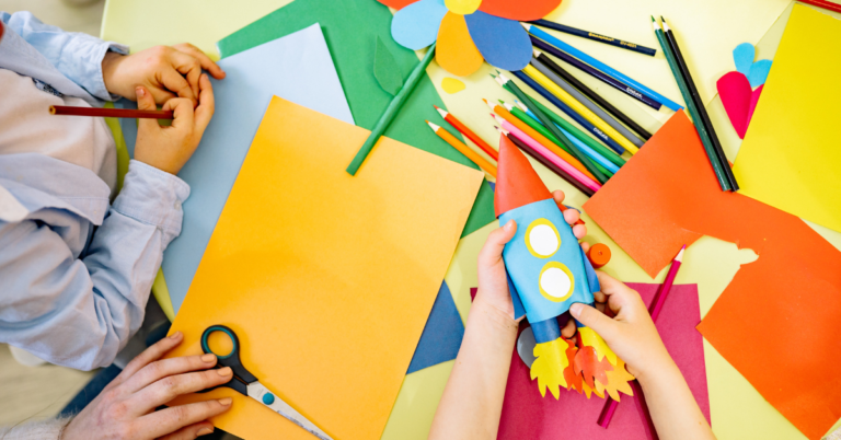 Two children using scissors, colored pencils, and craft paper at a table, the everyday school supplies that fine motor skills depend on